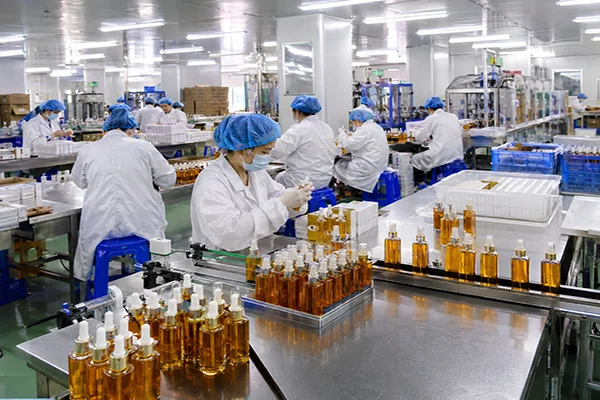 Рабочие в защитной одежде собирают и проверяют косметическую продукцию на производственной линии завода, на переднем плане — ряды флаконов.